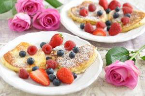 Photo of two plates of heart-shaped pancakes with fruit. Roses decorate the table.